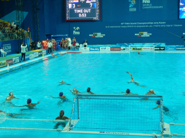 Eine Gruppe von Menschen spielt Wasserball in einem Pool während der 2015 FINA World Championships in Kazan, mit einem Netz in der Mitte, einer Menge im Hintergrund und Brettern mit Text, Logos, Trägern und einem Bildschirm mit Zahlen und Text.