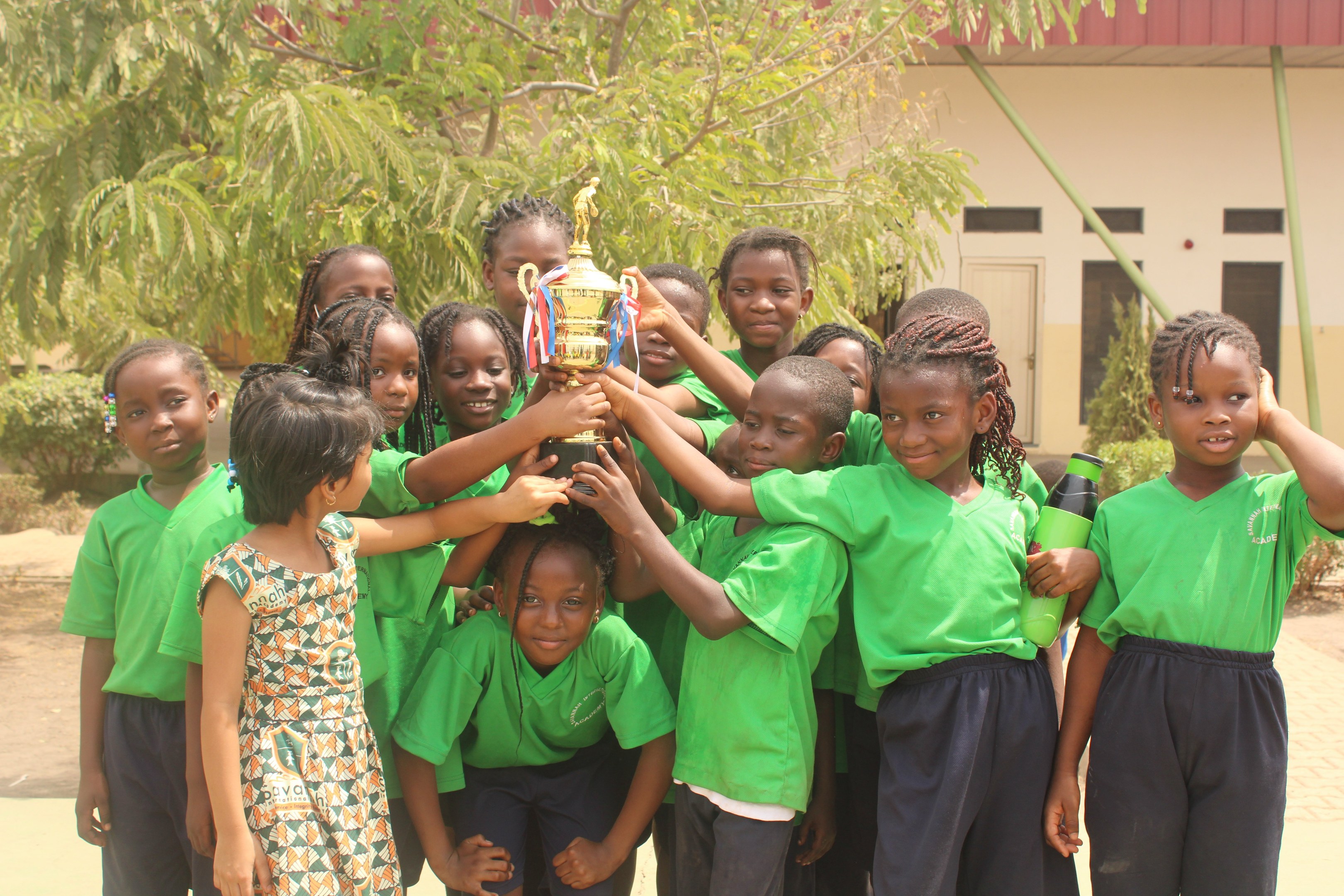 Gruppe von Kindern mit Pokal in einem Schulhof mit Bäumen, Pflanzen und einem Gebäude im Hintergrund