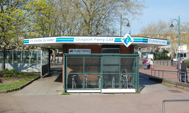Haltestelle mit einem 'Gosport Ferry Ltd'-Schild, Metallzaun, Menschen, Fahrrädern, Bäumen, Straßenlaternen, Schildern, einem Gebäude und einer bewölkten Himmel.