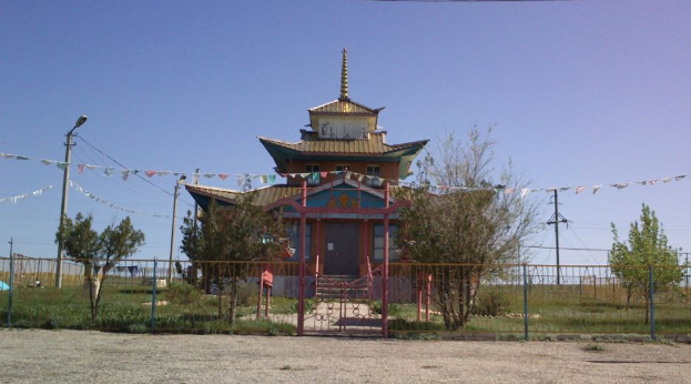 Ein kleines Gebäude mit einem Pavillon, das als Eingang zu einem tibetischen buddhistischen Tempel dient, steht in einem Schmutzfeld umgeben von einem Metallzaun, einem Tor, Pflanzen, Gras, Bäumen, Pfählen mit Drähten, einem Straßenpfahl und an Seilen befestigten Flaggen unter einem bewölkten Himmel.