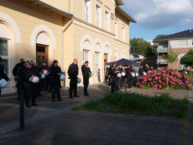Gruppe von Polizeibeamten vor einem Gebäude mit Fenstern und Türen, mit Helmen in der Hand, in der Nähe eines Zeltes, umgeben von Pflanzen, Gras, einem Laternenpfahl, Bäumen und einem bewölktem Himmel.