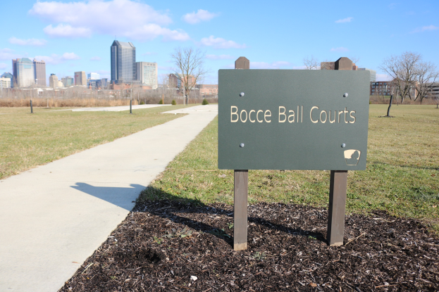 Ein Schild am Boden, das auf Boule-Plätze in Indianapolis, Indiana, hinweist, umgeben von Gras, einem Weg, Bäumen, Pfählen, einem Zaun, Gebäuden und einem bewölkten Himmel.