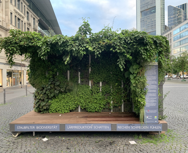 Grünes Gebäude mit Efeu an den Wänden, umgeben von Bäumen, Fahrzeugen, Strommasten und anderen Gebäuden unter einem bewölkten Himmel, Teil der Staufbilter Biodiversität in Berlin, Deutschland.