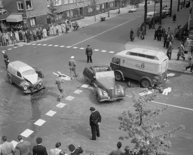 Schwarz-weiß-Szene eines Autounfalls an einer Straßenkreuzung mit mehreren Fahrzeugen, darunter ein Lieferwagen, und einer Gruppe von Zuschauern; Gebäude, Bäume, Strommasten und Schilder mit Text sind im Hintergrund zu sehen.