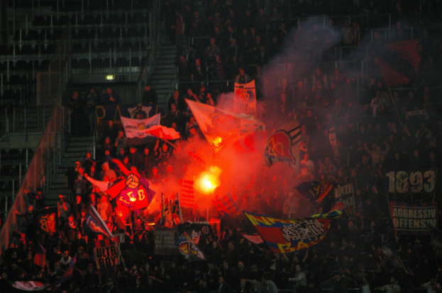 Eine große Menschenmenge in einem Stadion mit Fahnen und Bannern, beleuchtet von Lichtern, umgeben von Stufen und Geländern, mit einem Feuer in der Mitte.