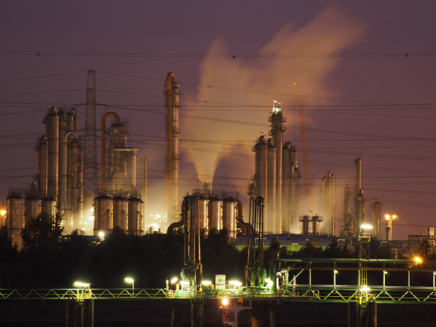 Ölraffinerie bei Nacht mit Rauchentwicklung, beleuchtet von umliegenden Lichtern mit sichtbarem Himmelhintergrund und Wasserzeichen.