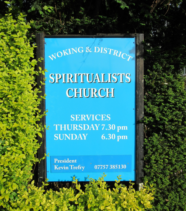 Ein hölzernes Brett mit der Aufschrift "Woking & District Spiritualists Church" im Freien, umgeben von Grün und einem Gebäude im Hintergrund.