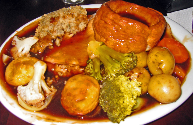 Ein weißer Teller mit Yorkshire-Pudding und einer Auswahl an Gemüse wie Karotten, Kartoffeln und Zwiebeln, das auf einem Tisch mit Gläsern, Papiertüchern und anderen Gegenständen drumherum steht.