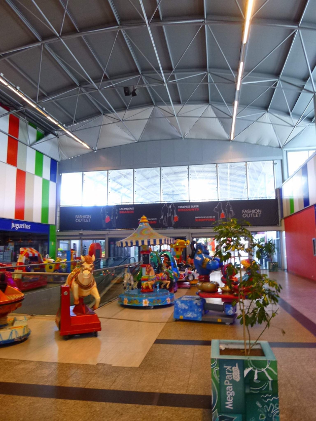 Amusement rides, a potted plant, scattered toys, informational boards, walls, ceiling lights, and metal poles in a large indoor shopping mall.