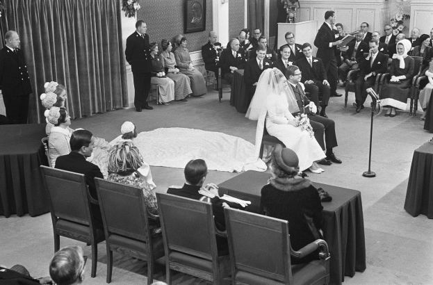 Schwarzes und weißes Foto einer Hochzeitszeremonie mit sitzenden und stehenden Menschen, einem Mann mit Mikrofon, einer Frau mit Blumen, einem Podium mit Mikrofon, Blumenschmuck, einem Vorhang, einem gerahmten Foto an der Wand und einer Tür im Hintergrund.