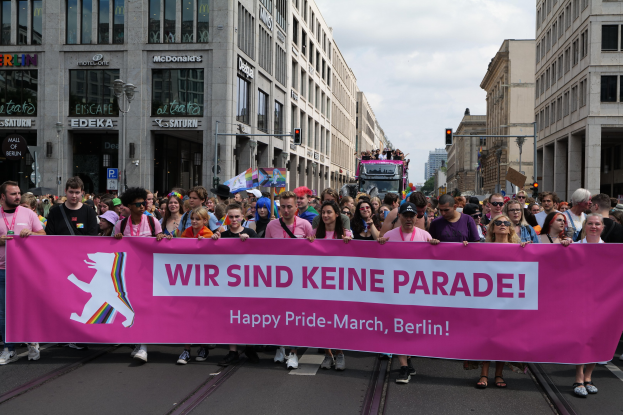 Eine Gruppe von Menschen, die eine pinkfarbene Fahne mit der Aufschrift "Happy Pride March" tragen, geht eine Straße in Berlin, Deutschland, entlang, mit Gebäuden, Laternen und Verkehrszeichen an der Straße unter einem bewölkten Himmel.