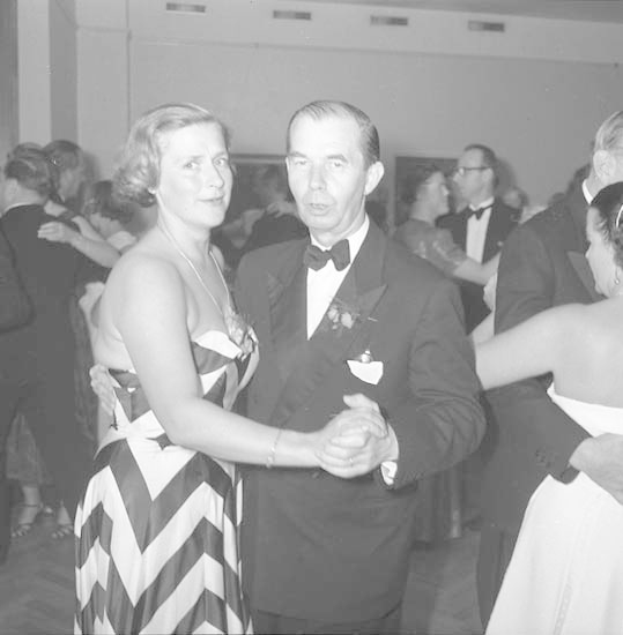 Ein Mann in einem schwarzen Blazer, weißem Hemd und schwarzen Hosen tanzt mit einer Frau in einem unbekannten Outfit auf einer formellen Veranstaltung, indem sie sich an den Händen halten und in der Nähe einer Wand zur Musik wiegen.