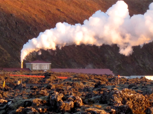 Ein Geothermiekraftwerk in Island mit Rauch aus seinem Schornstein, umgeben von Felsen im Vordergrund und Hügeln im Hintergrund.