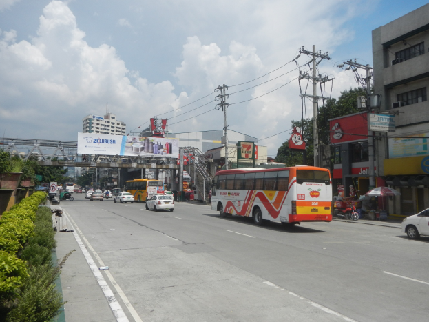 Eine Stadtstraße mit fahrenden Autos und Bussen, gesäumt von Gebäuden, Strommasten mit Drähten, Grünflächen, Schildern, einem Torbogen und einem bewölkten Himmel.
