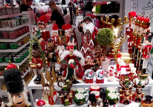 Ladeninnenausstattung mit Weihnachtsartikeln wie Spielzeug, Tassen und anderen Gegenständen auf Tischen und Regalen, mit Menschen, Lichtern und einer Wand im Hintergrund.