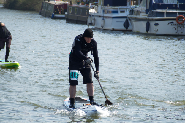 Zwei Personen stehen auf einem Brett im Wasser und paddeln; im Hintergrund sind Boote und in der Ferne Täler zu sehen; die Person in der Mitte steht auf einem Surfbrett und hält ein Paddel.