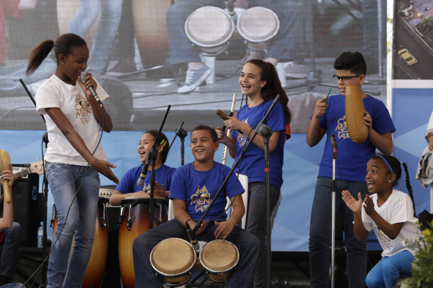 Kinder, die auf einer Bühne Musikinstrumente spielen, mit einer Frau, die ein Mikrofon hält, umgeben von Mikrofonständern und einem Banner im Hintergrund, mit Pflanzen auf der rechten Seite.