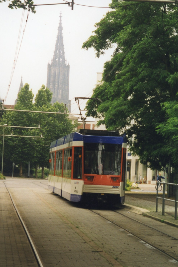 Rotes und weißes Zugfahrzeug auf Schienen neben einem hohen Gebäude, mit einer Person, die auf einem Gehweg rechts neben den Schienen Fahrrad fährt, Bäume entlang der Schienen und Gebäude im Hintergrund unter einem klaren blauen Himmel.