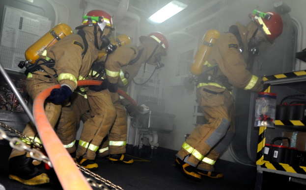 Eine Gruppe von Feuerwehrleuten in Schutzausrüstung, darunter Helme und Sauerstoffflaschen, arbeitet an einem Feuerwehrauto, mit einem Gestell voller verschiedener Gegenstände auf der rechten Seite und Rohren, Lichtern und anderen Gegenständen im Hintergrund.