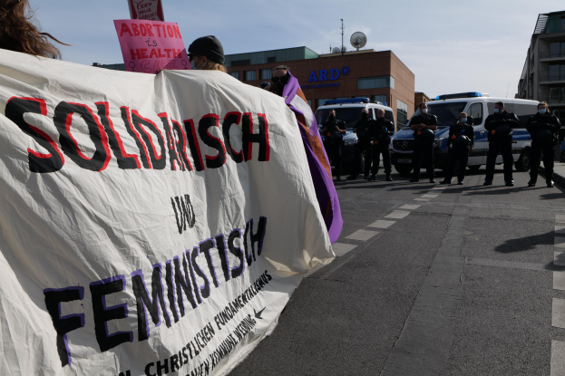 Eine Gruppe von Menschen marschiert die Straße entlang und hält ein Banner mit der Aufschrift "Solidarität und Feminismus", mit parkenden Fahrzeugen und Gebäuden im Hintergrund unter einem klaren blauen Himmel.