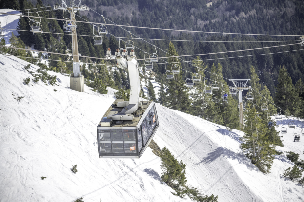 Eine volle Seilbahn fährt einen schneebedeckten Berg hinauf, mit hohen Bäumen im Hintergrund.