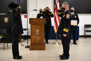 Gruppe uniformierter Personen in Masken bei einer Vereidigung eines neuen Feuerwehrchefs, der eine Urkunde hält, mit Fahnen, Podium, Stühlen, Tischen und einer Wand im Hintergrund.