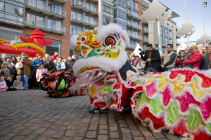 Ein lebhaftes chinesisches Neujahrsfest in Amsterdam mit einem Löwen tanzen vor einem Publikum, das Fotos macht, vor einer Kulisse aus Gebäuden, Laternenmasten und einem klaren blauen Himmel.