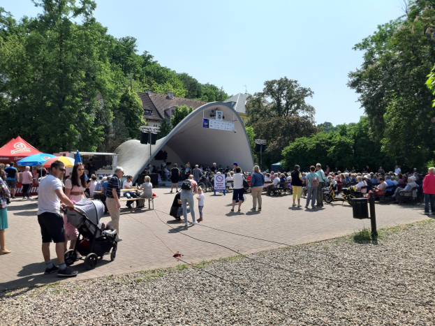 Eine Menschenmenge versammelt sich vor einem Gebäude bei der Festival-Eröffnung, einige sitzen auf Bänken, andere stehen; ein Kinderwagen ist im Vordergrund, Bäume und Gebäude säumen den Hintergrund unter einem klaren blauen Himmel, Zelte sind links und ein Mülleimer rechts.