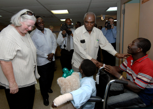 Eine Gruppe von Menschen mit Kameras umgibt ein Kind im Rollstuhl, das ein weißes Hemd trägt und einen Verband am Arm hat, in einem Raum mit Deckenlampen und einer Tür im Hintergrund.