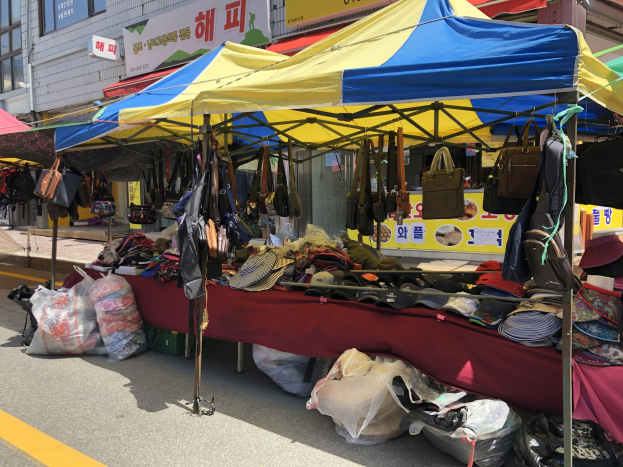 Ein Straßenmarkt mit Zelten, die verschiedene Gegenstände wie Taschen und Hüte präsentieren, vor Häusern mit Fenstern und Texttafeln.