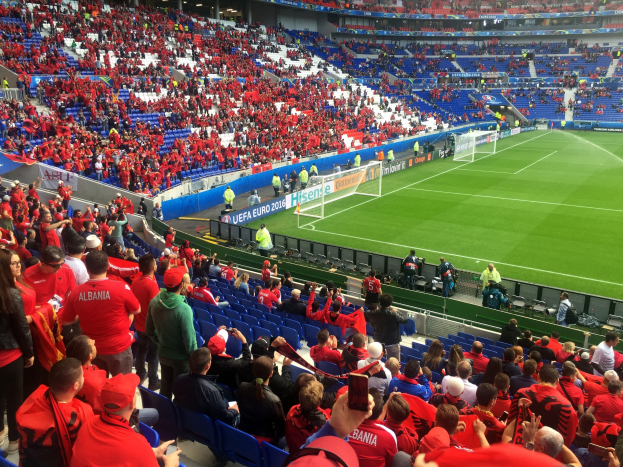 Eine große Menschenmenge in einem Stadion schaut einem Fussballspiel zu, mit Menschen, die auf Stühlen sitzen und stehen, Bannern, einem Torpfosten und Deckenleuchten.