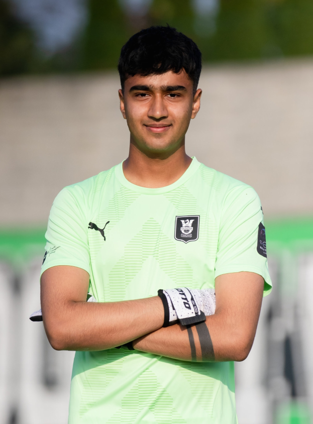 Ein junger Mann in einem grünen T-Shirt steht auf einem Fußballfeld mit verschränkten Armen und lächelt, mit einem unscharfen Hintergrund.