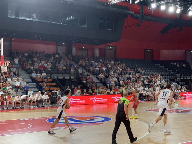 Eine Gruppe von Menschen, die Basketball in einer Turnhalle spielt, mit einem Basketballkorb auf der linken Seite und Zuschauern im Hintergrund, unter hellen Stadionlichtern.