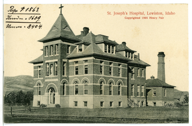 Ein Schwarz-Weiß-Foto des St. Joseph-Krankenhauses in Lewiston, Idaho, das ein großes Gebäude mit Fenstern, eine Treppe mit Geländern, einen Schornstein auf der rechten Seite und Text oben zeigt, mit Bäumen, Hügeln und Himmel im Hintergrund.