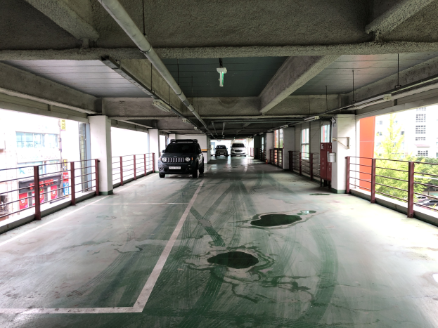 Ein mehrgeschossiges Parkhaus mit Autos zwischen Geländern, Säulen, Lichtern und Rohren, umgeben von Bäumen und gegen den Hintergrund von Gebäuden mit Fenstern in einer Stadtlandschaft.