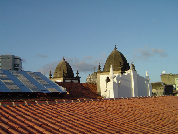Stadtansicht mit Gebäuden im Vordergrund, Solarpanels auf einem Dach und einem blauen Himmel im Hintergrund, was den Einsatz von erneuerbarer Energie anzeigt.