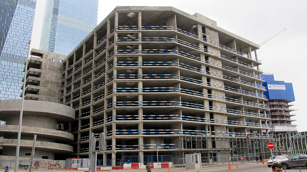 Großes Bauprojekt in einer von Hochhäusern umgebenen Stadt mit Fahrzeugen, Fußgängern, Verkehrsinfrastruktur und Kränen vor einem klaren blauen Himmel.