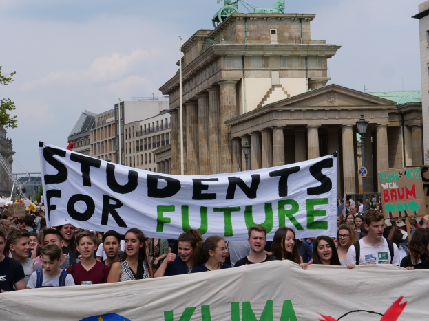 Eine Gruppe von Schülern marschiert in Berlin, hält ein buntes Banner mit der Aufschrift "Schüler für die Zukunft" und hat Gebäude, Bäume und Himmel im Hintergrund.