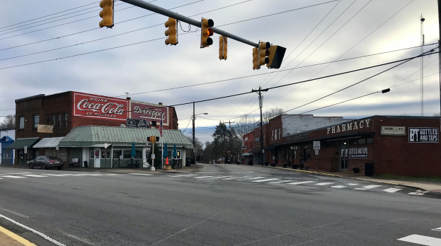 Eine Stadtstraßenkreuzung mit Verkehrsampel, Fahrzeugen, Gebäuden mit Namensschildern, Strommasten mit Drähten, Bäumen und einem bewölkten Himmel.
