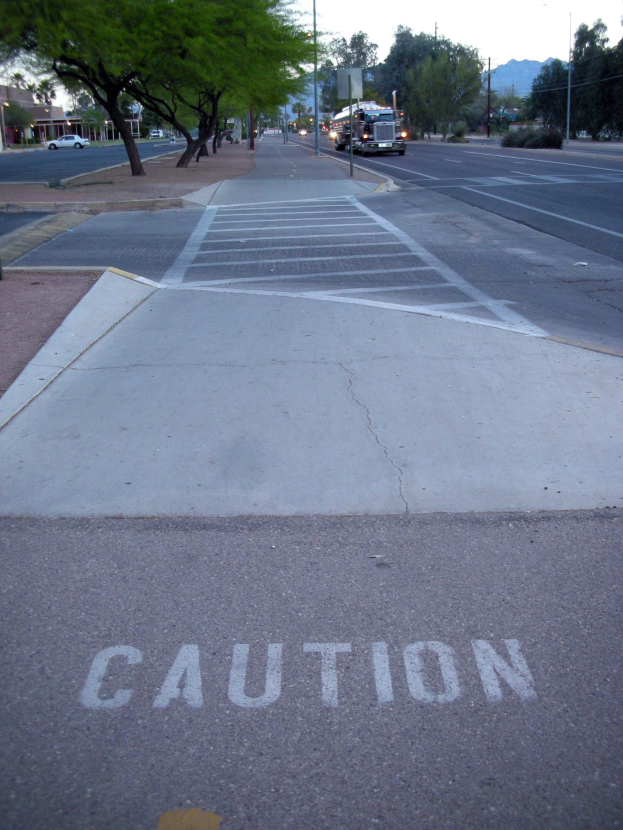 Stadtstraße mit fahrenden Fahrzeugen, ein Gehweg mit einer Warnschilder neben einem Baum, Strommasten, Straßenlaternen, Bauzäune, Grünfläche, Gebäude, entfernte Berge und einen klaren blauen Himmel.