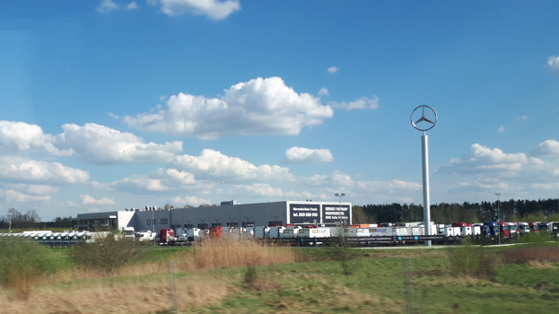 Mercedes-Benz-Fabrik in Deutschland mit Fahrzeugen auf der Straße, Grünflächen, Gebäuden und einem bewölkten Himmel.
