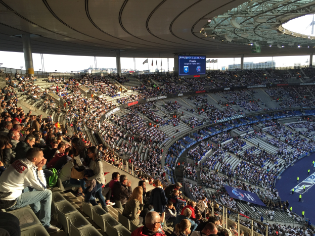 Große Menschenmenge in einem Stadion bei einem Fötballspiel, mit einer Bühne rechts, Fahnen, Masten, einem Bildschirm und dem Allianz Stadion in München, Deutschland im Hintergrund.