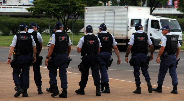 Gruppe von Beamten in Uniform auf einer Straße mit einem geparkten Lkw, Gebäuden, Bäumen und einer Brücke im Hintergrund.
