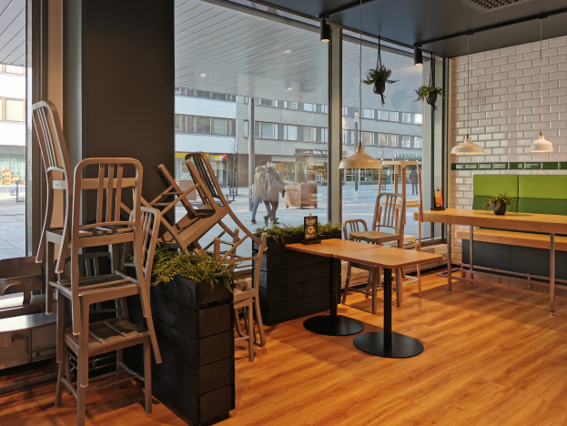 Ein Restaurant in Berlin, Deutschland, mit Holztischen und -stühlen vor einem großen Fenster, das Menschen auf der Straße, Gebäude und den Himmel zeigt, umgeben von Pflanzen und Lichtern.